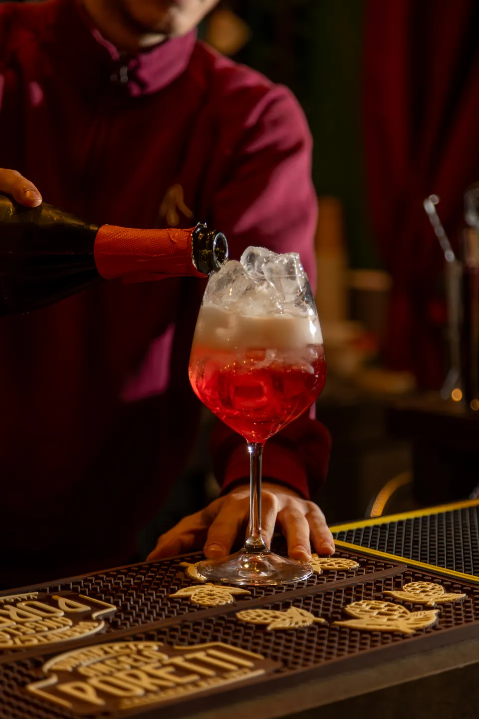 Un calice con ghiaccio e un drink rosso, completato versando da una bottiglia, sul tappetino del bancone del bar
