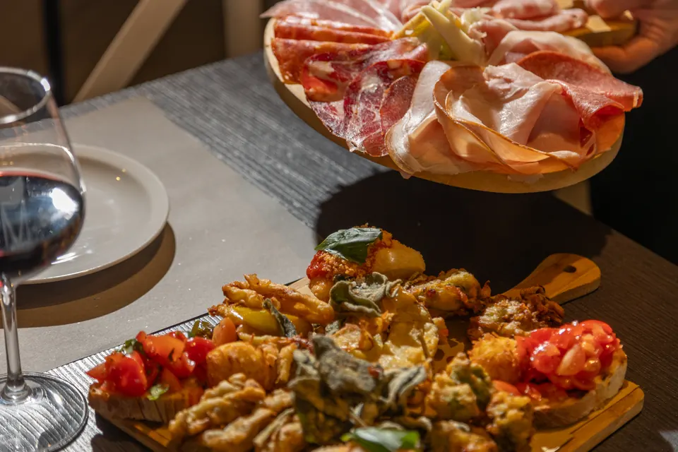 Un tagliere di salumi, fritti e bruschette con pomodoro, accanto a un bicchiere di vino rosso