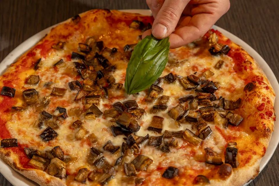 Una pizza con melanzane a cubetti, mentre una mano aggiunge una foglia di basilico